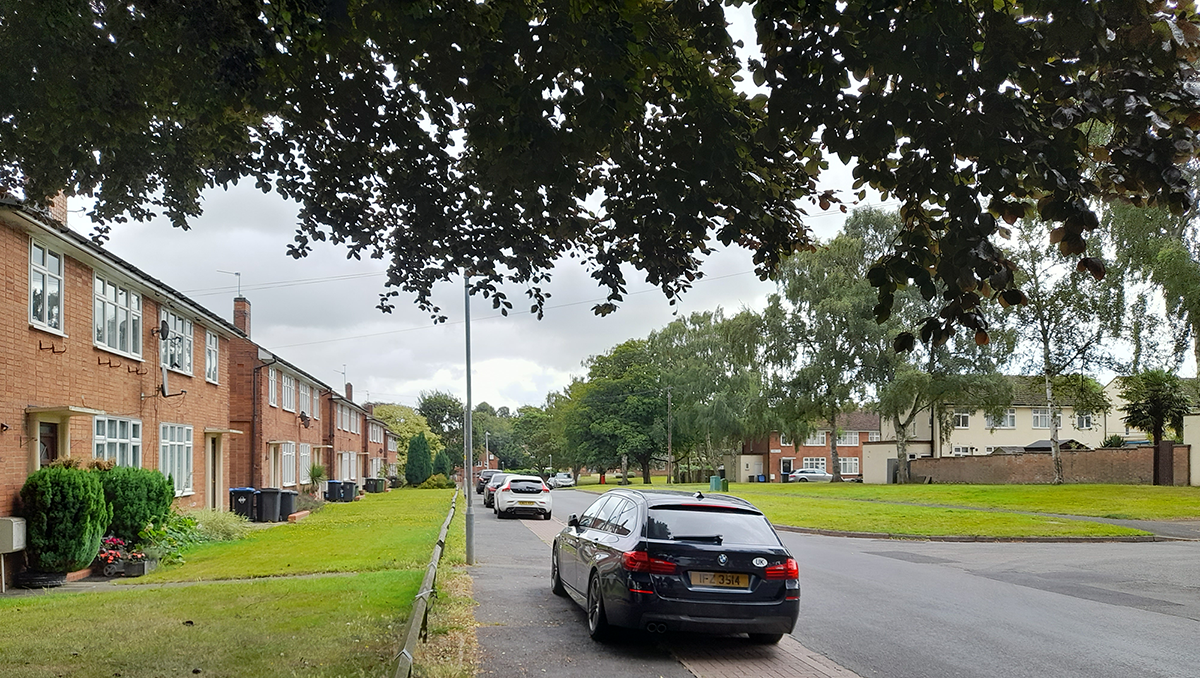 A quiet, tree-lined residential street in the Bilton estate featuring red-brick terrace houses, parked cars, and a prominent red graphic overlay.