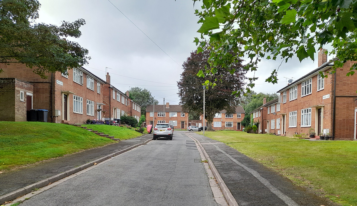 A street view of the existing Bilton housing estate, showing red-brick maisonettes lining a quiet residential road with grass verges and mature trees.